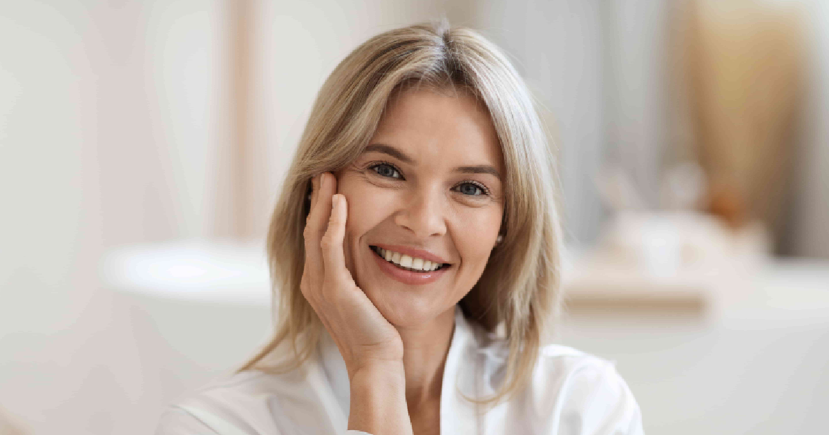 Smiling middle-aged woman in a white robe touching her face, showcasing healthy, youthful skin and natural beauty indoors after Peptide Therapy in Cedar Park, TX.